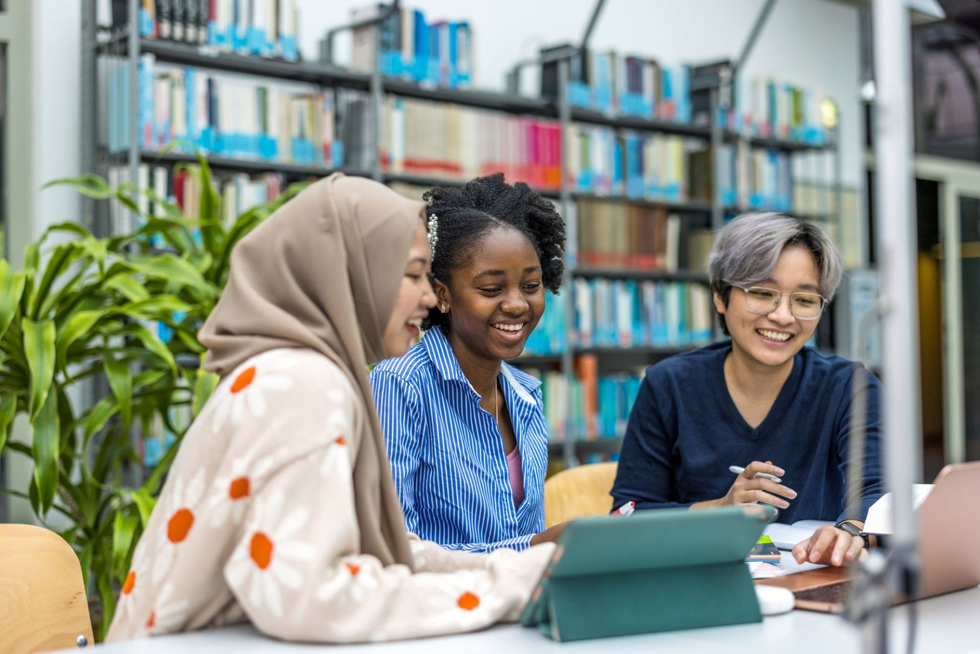 Three people collaborating at a table in a library with bookshelves and greenery in the background.