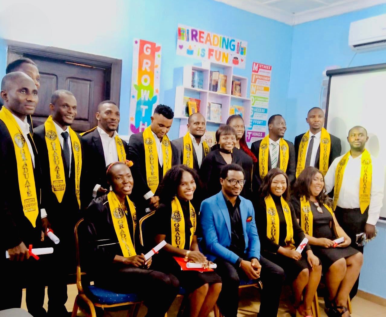 Group of graduates in yellow sashes posing in a room with colorful educational posters and books.