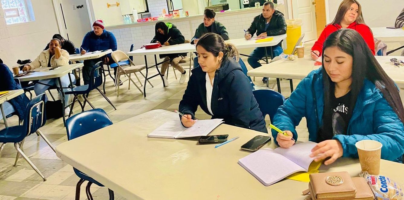 Adults seated at tables in a classroom setting, taking notes and focused on their work.